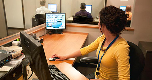 Test center proctor observing candidates testing both through the enclosed glass proctor station and on the computer streaming live digital video.